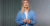 thumb-Iryna Remihailo stands in front of a gray wall with a geometric pattern. She is wearing a light shirt over a white T-shirt and holding her hands in front of her.