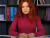 thumb-Nataliia Ostapiuk, with red hair, sits at a table wearing a pink sweater and has a serious expression. On the table are a pen, calculator, and papers. In the background, there are bookshelves with folders of various colors