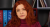 thumb-Nataliia Ostapiuk, with red hair, sits in a pink sweater and has a serious expression. In the background, there are bookshelves with folders of various colors