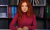 thumb-Nataliia Ostapiuk, with red hair, sits in a pink sweater and has a serious expression. In the background, there are bookshelves with folders of various colors